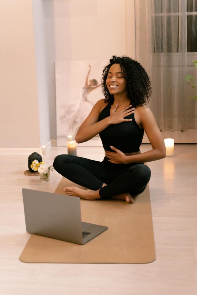 Image of a woman meditating indoors on a yoga mat, smiling with one hand on her chest and candles nearby, showing postpartum belly workouts.