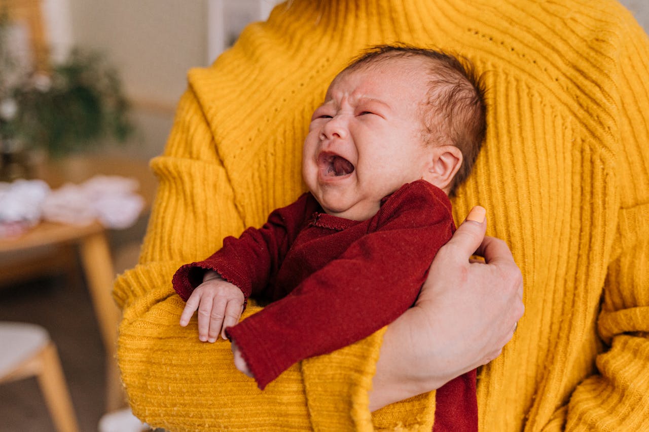 Close-up image of a crying baby wearing a red outfit being held by a person in a bright yellow sweater, representing breastfeeding mother eat to prevent colic.