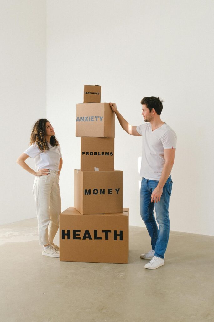 Image of a man and a woman stacking cardboard boxes labeled health, money, problems, anxiety, and happiness.