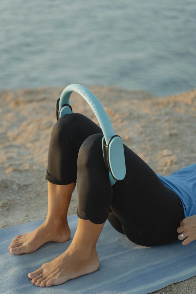 Image of a woman lying on a yoga mat by the sea, using a fitness ring for leg exercises.