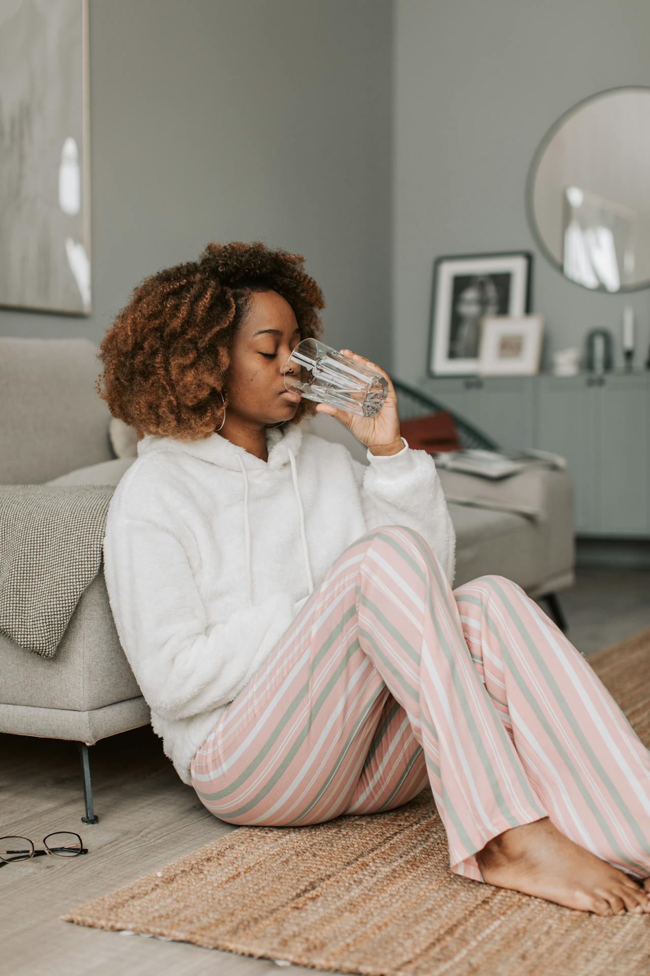 Image of a woman in cozy striped pajama pants and a hoodie sitting on the floor, drinking a glass of water, representing anti-colic diet for breastfeeding mothers.