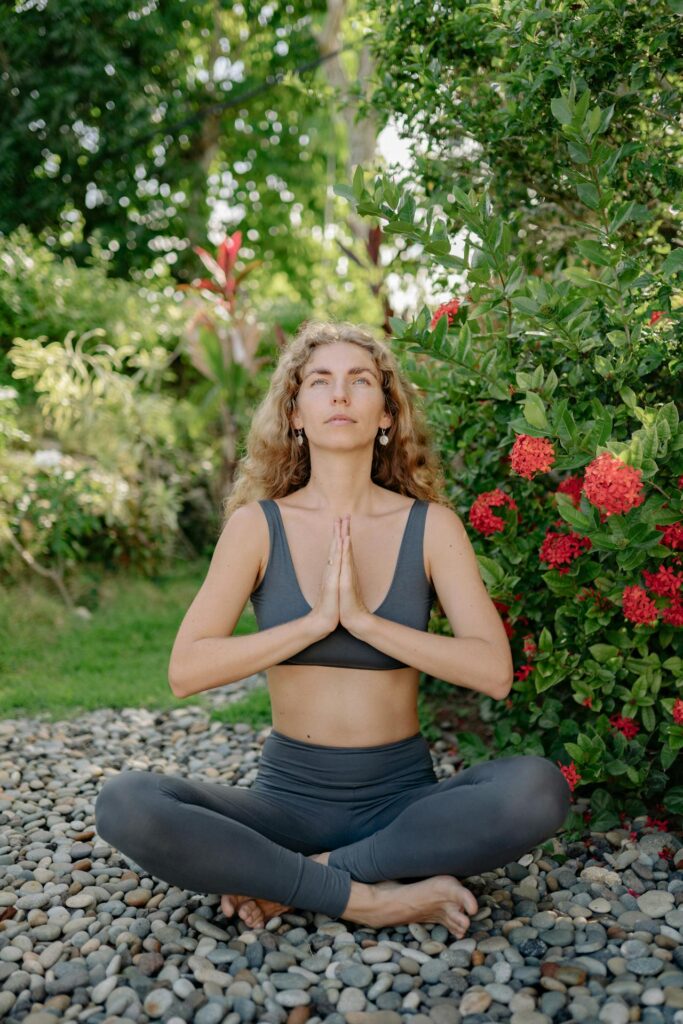 Close-up image of a woman sitting cross-legged outdoors, meditating with palms together in a serene garden.