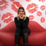 Image of a woman in black sits on a red lips-shaped couch against a wall with red lipstick kiss prints, showing what a women really want for valentines.