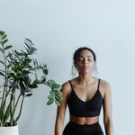 Close-up image of a woman in black workout clothes sits on a wooden bench with closed eyes, meditating near a plant, representing how to prioritize self-care as a mom.