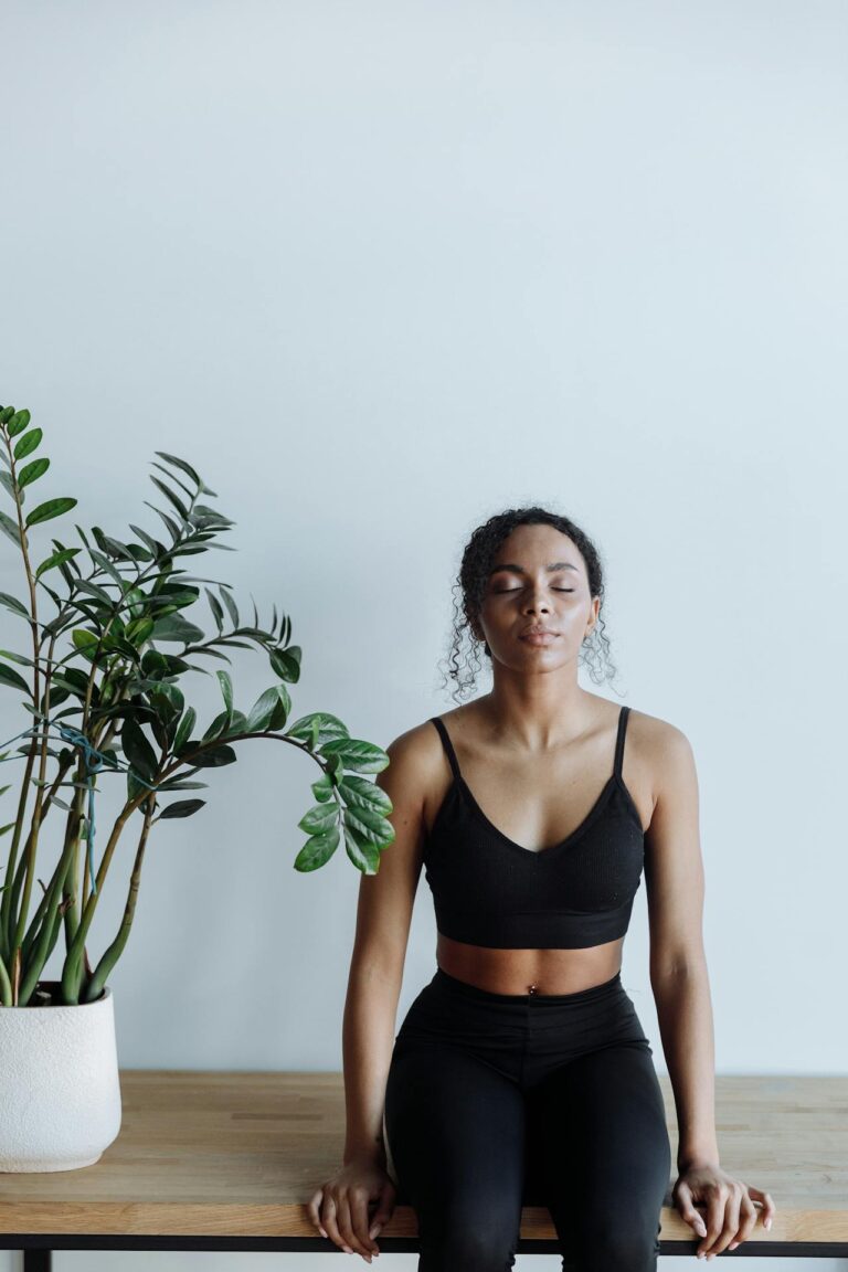 Close-up image of a woman in black workout clothes sits on a wooden bench with closed eyes, meditating near a plant, representing how to prioritize self-care as a mom.