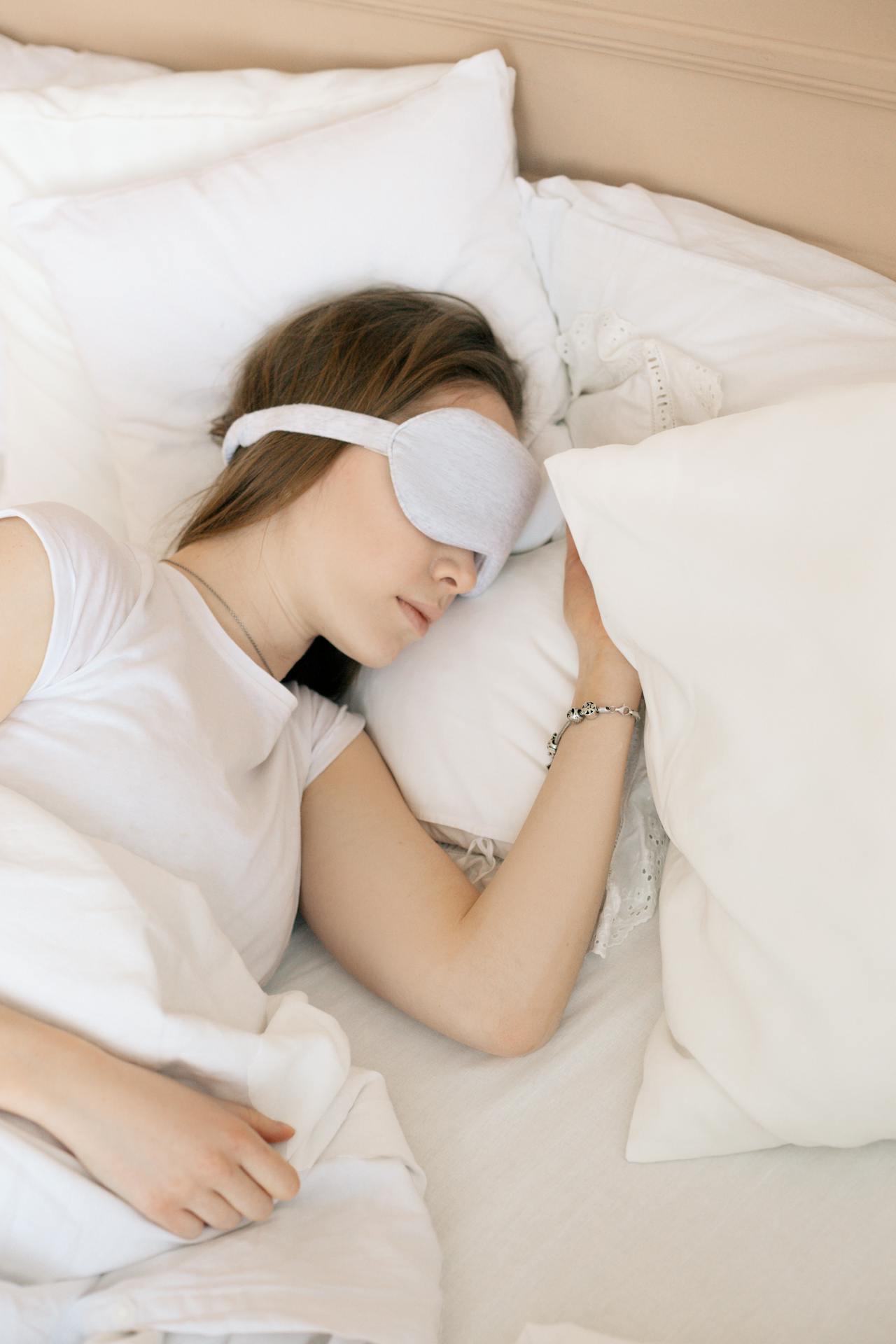 Image of a woman in a white shirt sleeps on a bed with white pillows and sheets, wearing a gray sleep mask, representing thoughtful Valentine’s gifts for her.