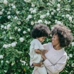 Image of a mother holding her laughing child in front of a bush with white flowers, both smiling joyfully, showing conscious parenting vs gentle parenting.