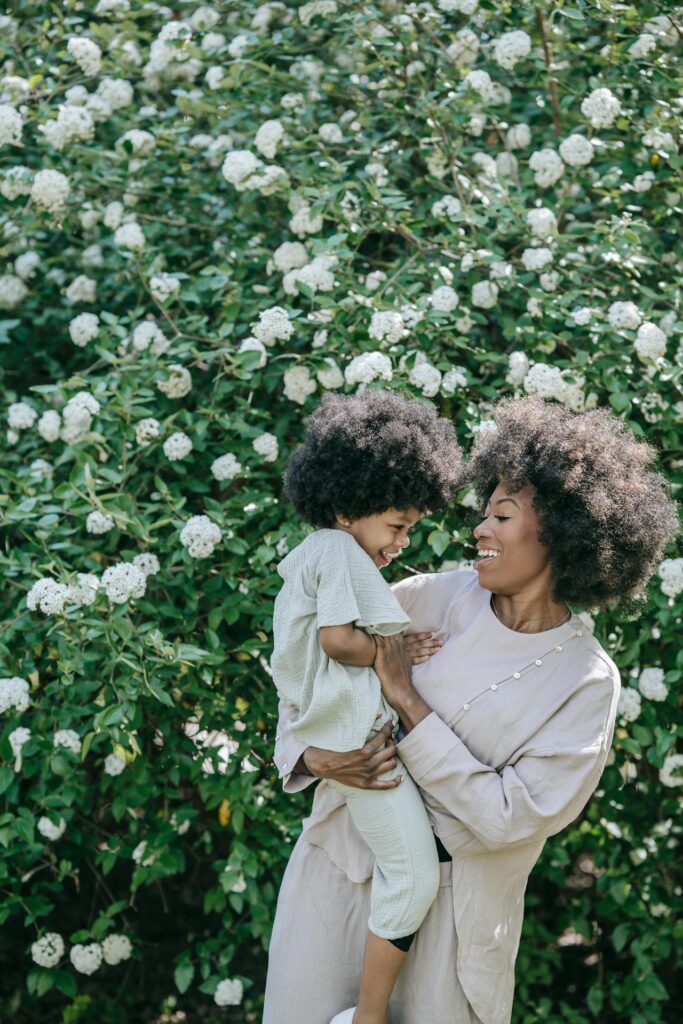 Image of a mother holding her laughing child in front of a bush with white flowers, both smiling joyfully, showing conscious parenting vs gentle parenting.