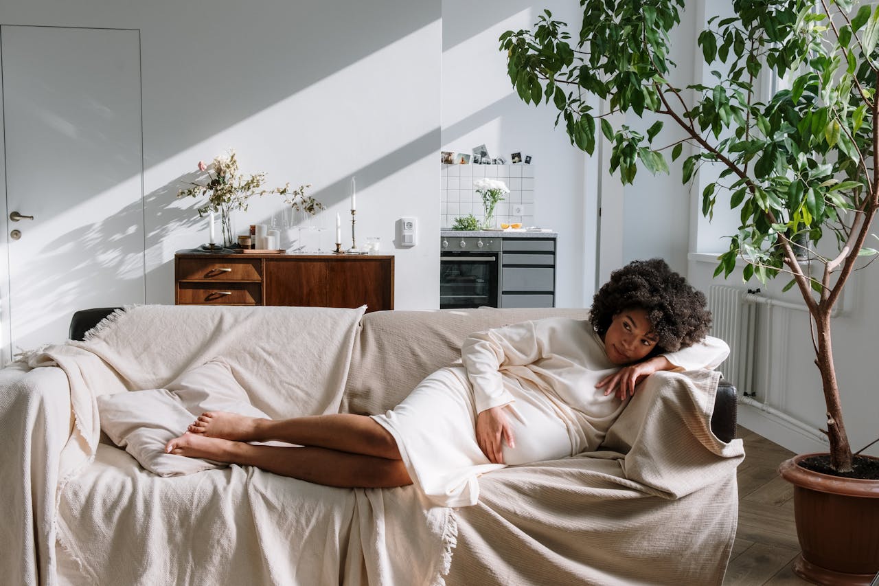 Image of a pregnant woman in a cream dress reclining on a beige couch in a sunlit living room with plants.