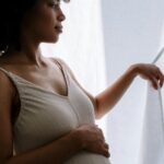 Image of a pregnant woman in a beige dress gently holds her belly while gazing out of a sunlit window, showing weird signs labor is near.