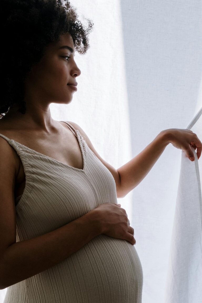 Image of a pregnant woman in a beige dress gently holds her belly while gazing out of a sunlit window, showing weird signs labor is near.