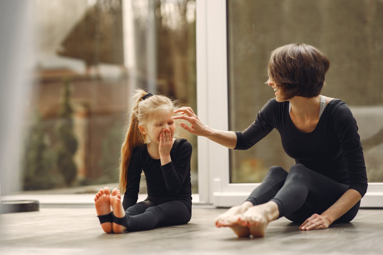 Image of a mother and her daughter wearing black outfits sit on the floor, smiling and bonding during exercise, representing similarities between conscious parenting and gentle parenting.