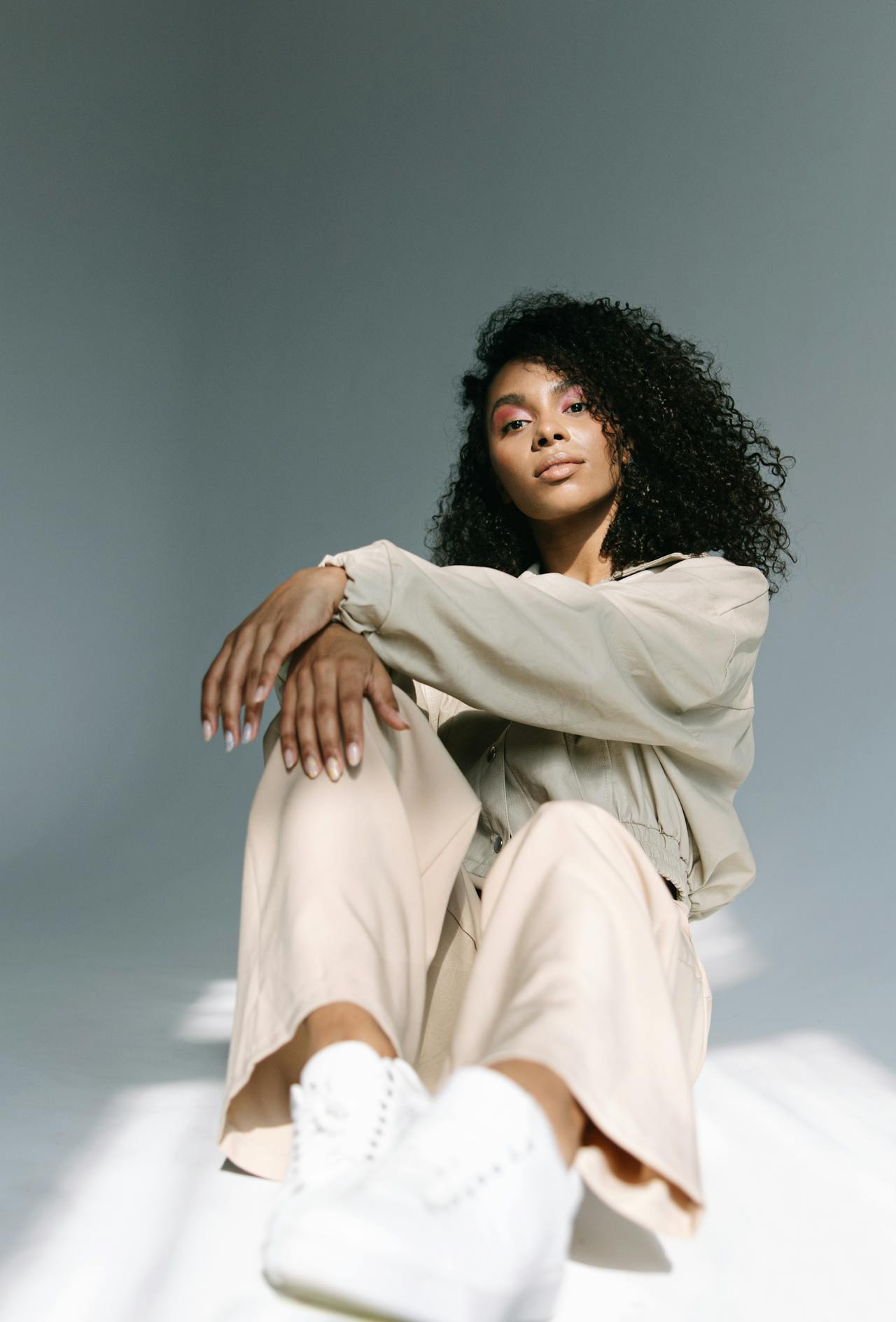 Image of a woman with curly hair seated on the floor, wearing a beige jacket and white sneakers, showing ow to dress stylishly with kids.