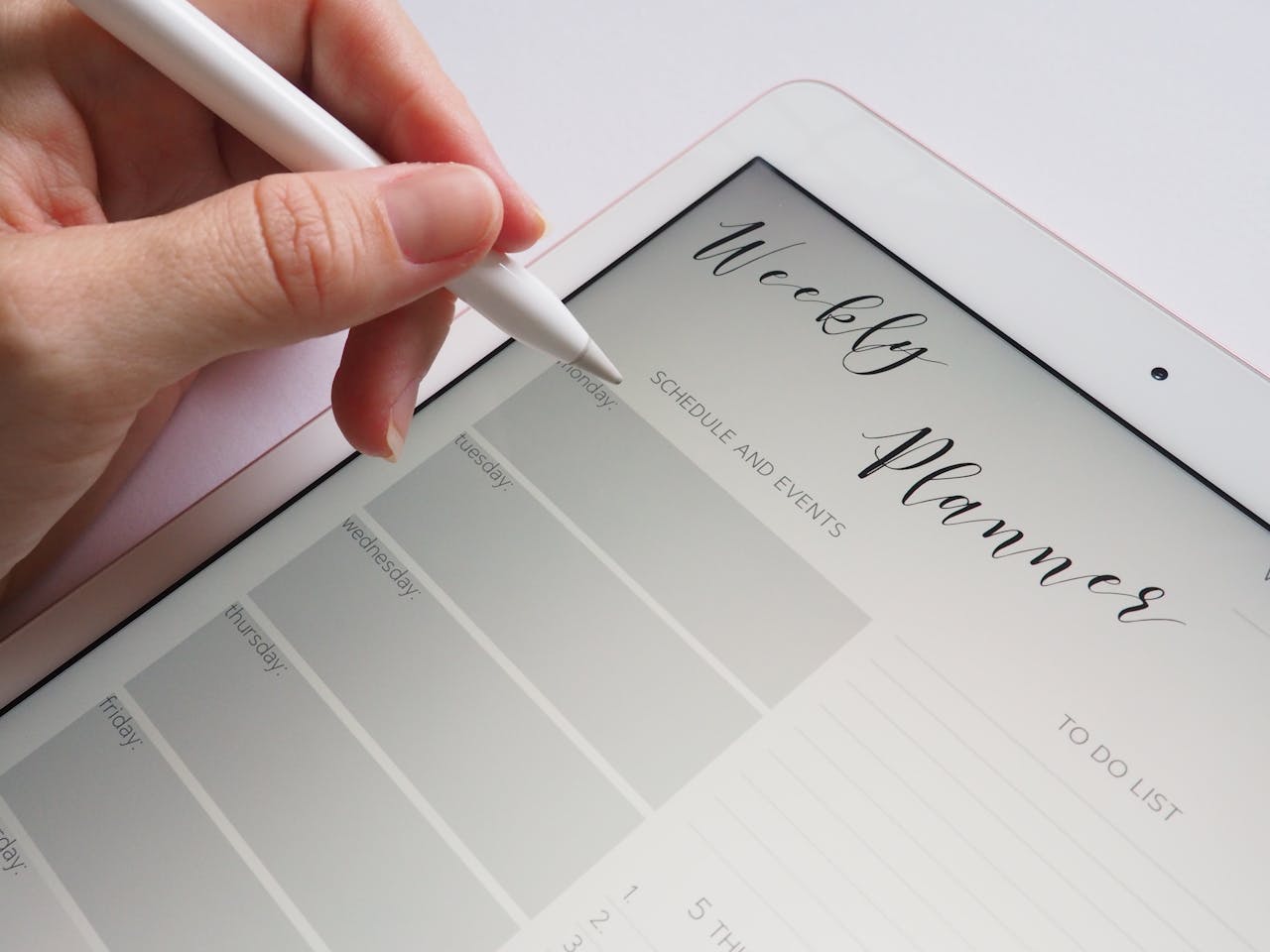 Close-up image of a hand holding a stylus pen, writing on a digital weekly planner displayed on a tablet.