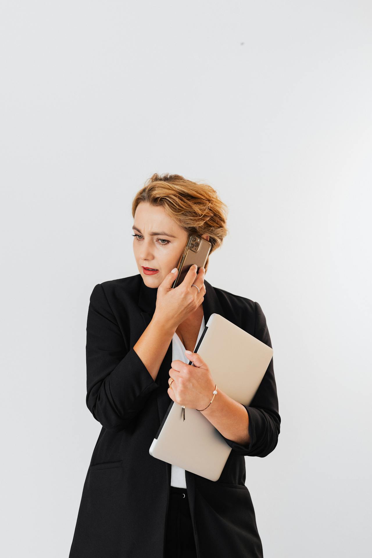 Image of a stressed woman in a black blazer holds a laptop and talks on the phone with a concerned expression.