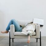 Image of a woman is slumped on a beige armchair with a notebook covering their face, appearing exhausted, showing signs you aren't practicing self-care as much as you should be.