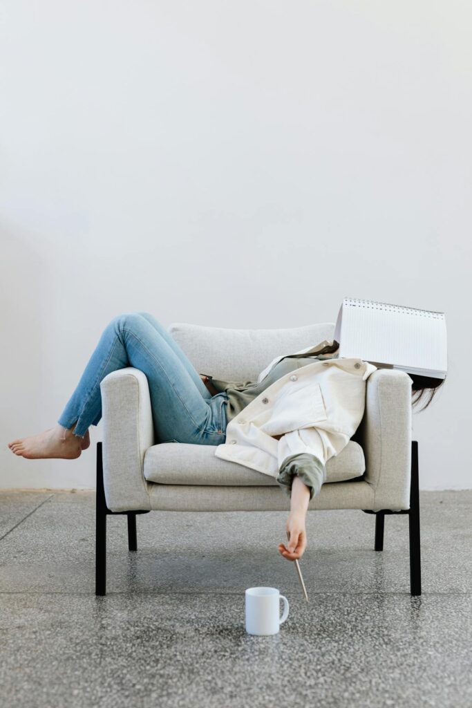 Image of a woman is slumped on a beige armchair with a notebook covering their face, appearing exhausted, showing signs you aren't practicing self-care as much as you should be.