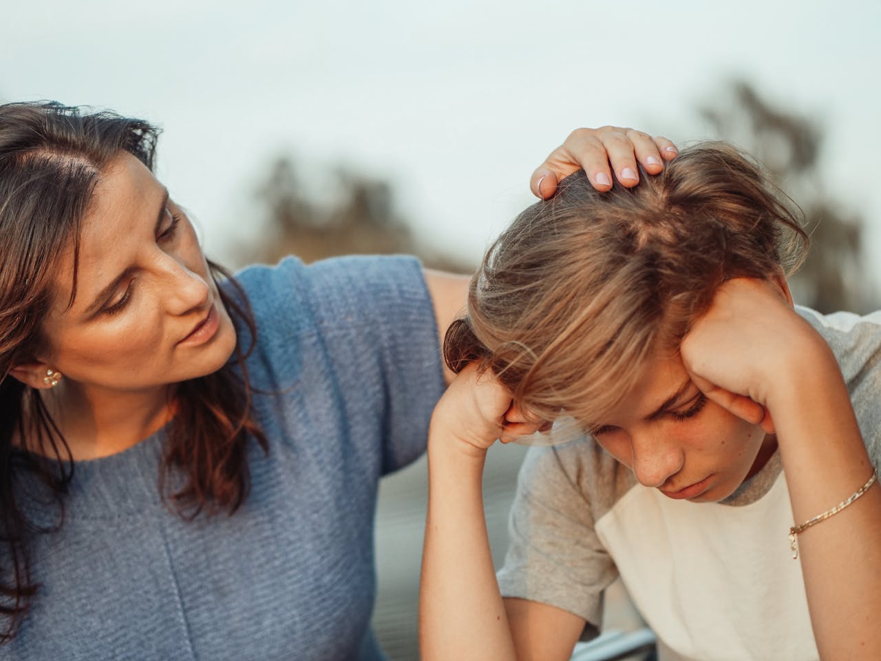 Image of a mother comforting her sad son, placing her hand on his head as he rests his face in his hands, showing conscious parenting vs gentle parenting.