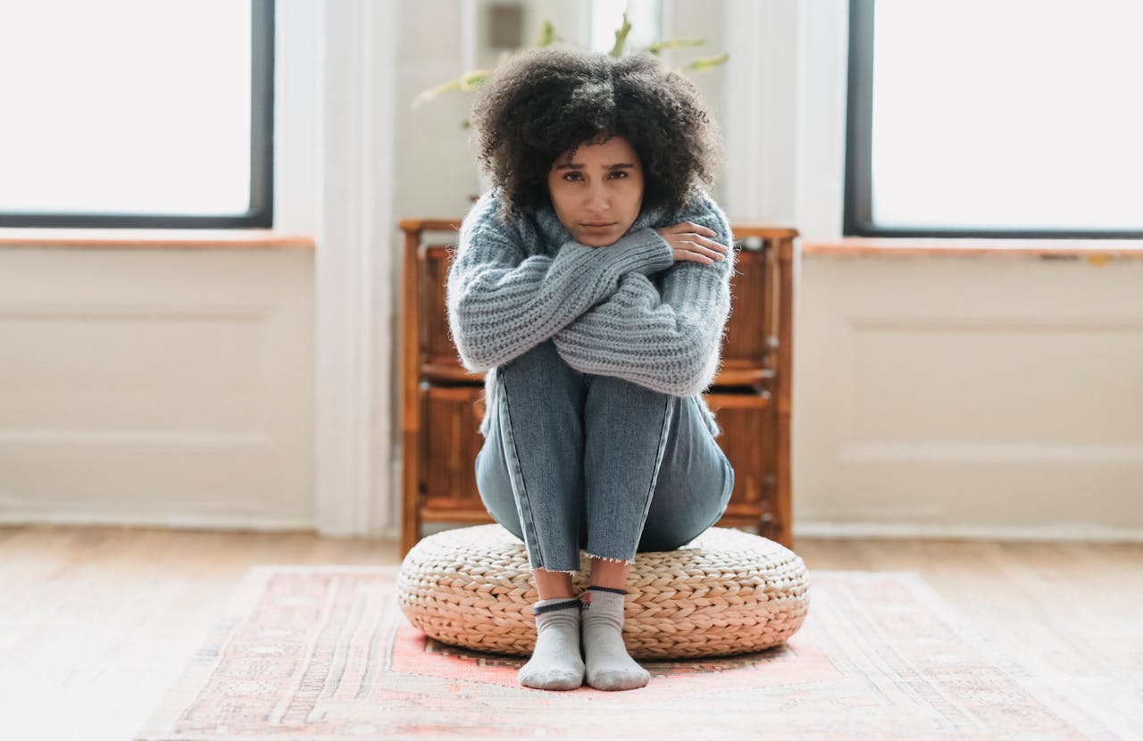 Image of a woman sits on a pouf, hugging herself tightly in a cozy sweater, looking cold or distressed, representing signs you aren’t practicing self-care as much as you should be.