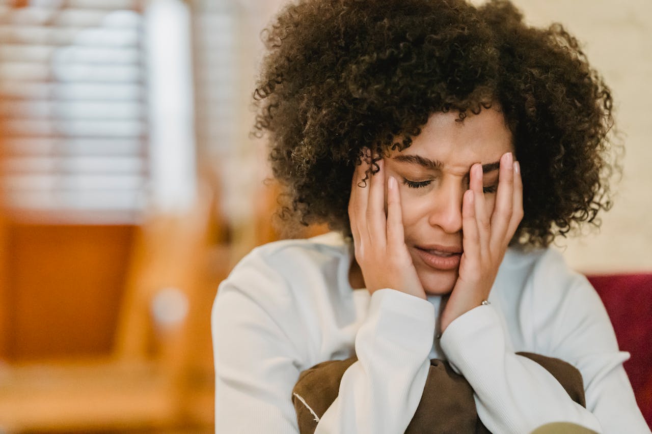 Image of a distressed woman with curly hair sits indoors, holding her face in her hands, appearing sad or overwhelmed.