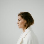 Image of a woman in a white coat with neatly styled hair looks to the side against a plain light gray background, showing how can moms stay stylish and comfortable daily.