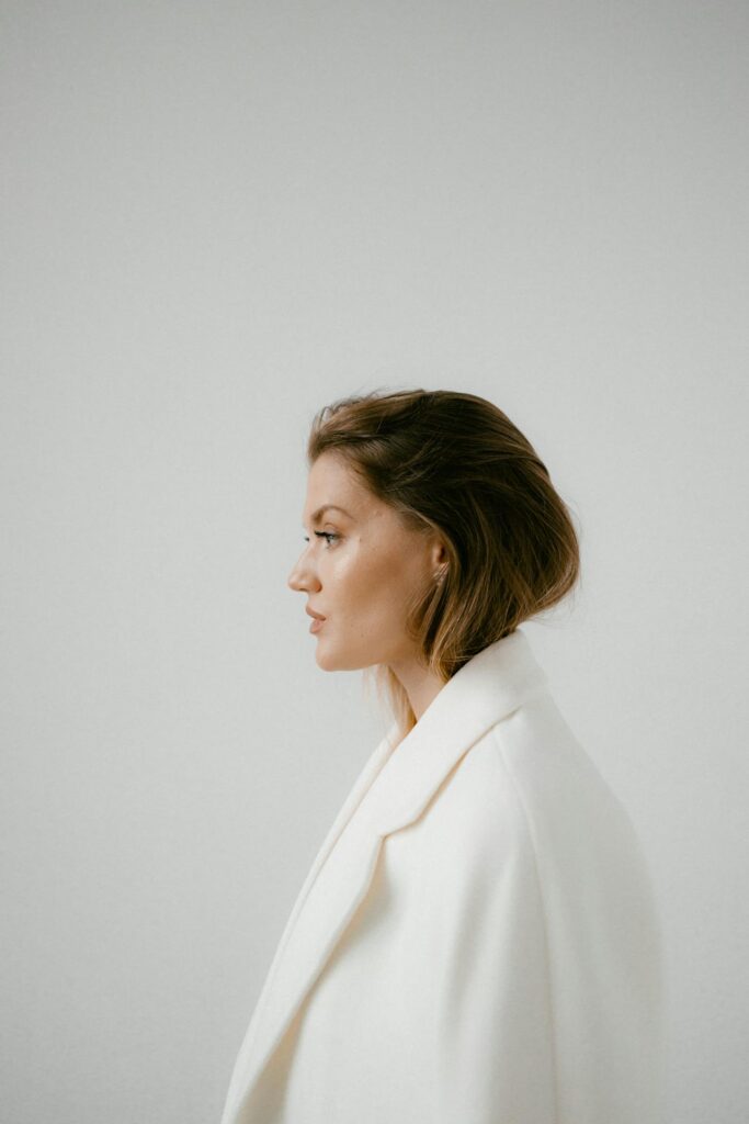 Image of a woman in a white coat with neatly styled hair looks to the side against a plain light gray background, showing how can moms stay stylish and comfortable daily.