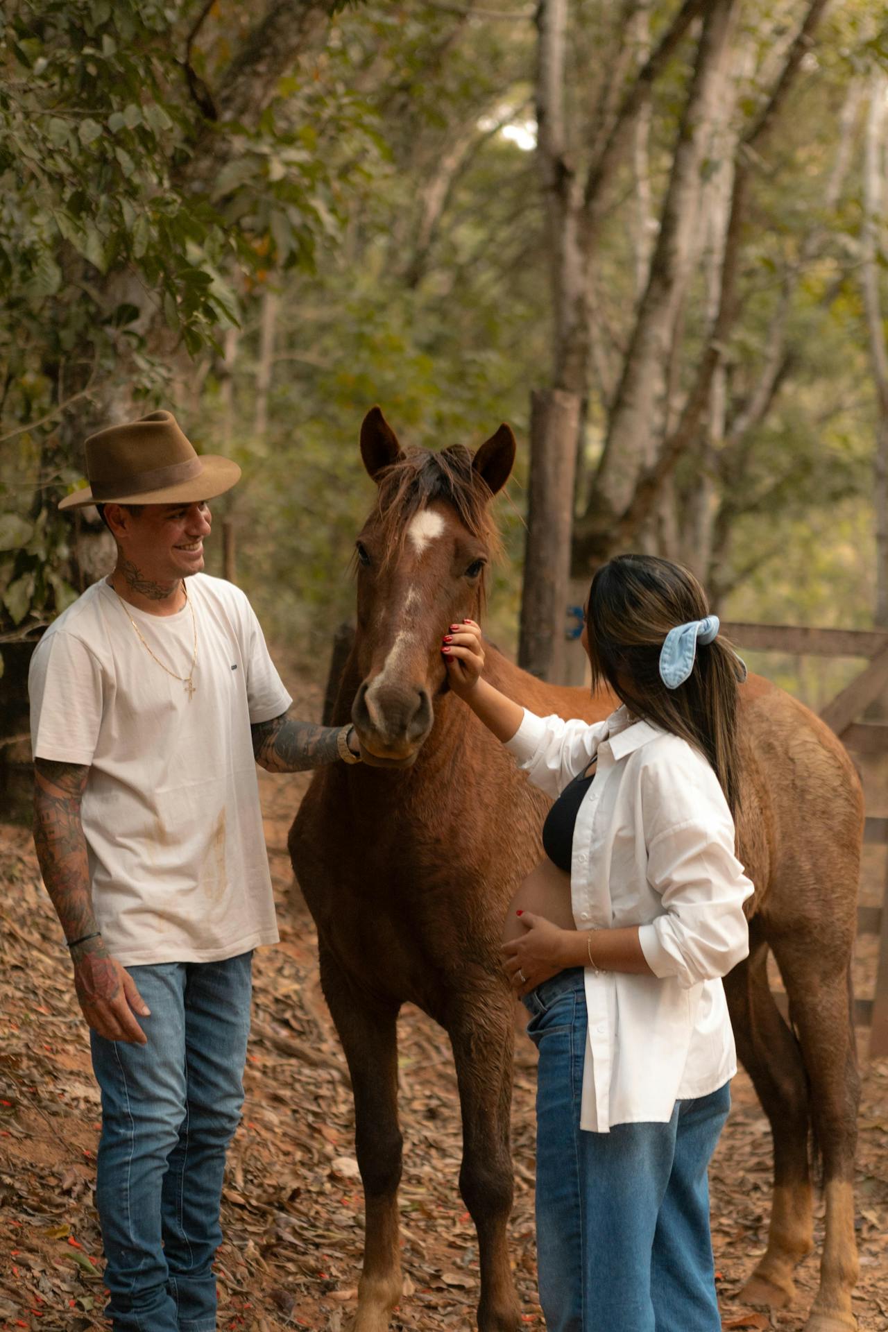 Image of a couple pets a brown horse in a forest setting, with the woman wearing a blue hair tie and white shirt.