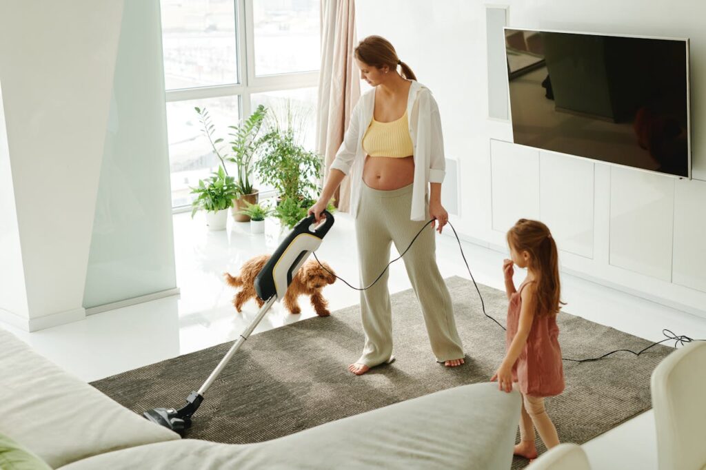 Image of a pregnant woman vacuums a carpet while her daughter watches and a small dog plays nearby in a bright room, showing emotional signs labor is approaching.