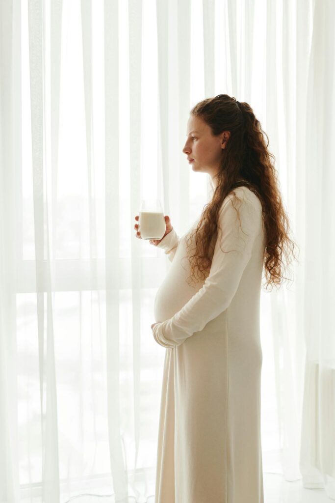 Image of a pregnant woman in a long dress stands by a window, holding a glass of milk and gazing outside.