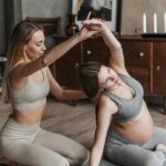 Image of a pregnant woman in gray activewear does a side stretch on a yoga mat, assisted by a trainer, showing how to turn prodromal labor Into real labor.