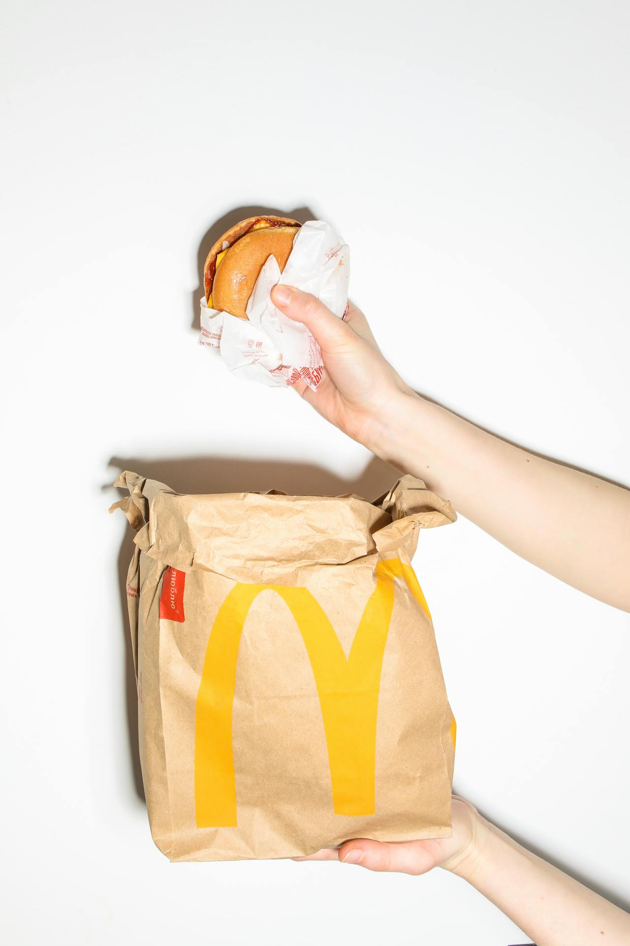 Image of a hand holds a McDonald's burger, with another hand holding a McDonald's paper bag against a white background.