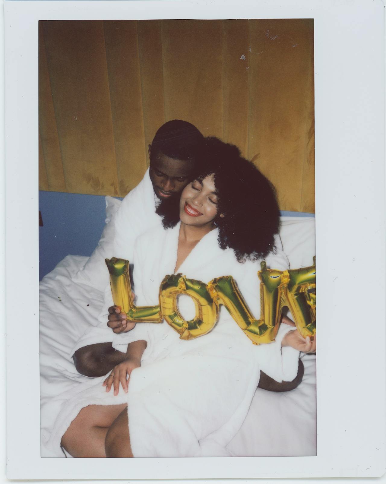 Image of a couple in white robes on a bed holds gold balloon letters spelling LOVE, smiling and embracing, showing what do women really want for valentines.