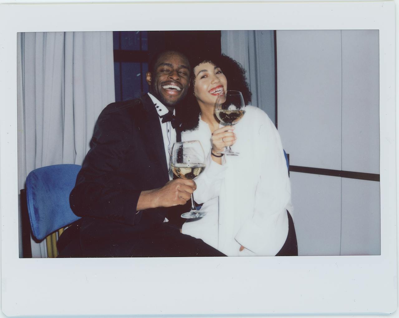 Close-up image of a happy couple dressed elegantly smiles while holding wine glasses, captured in a Polaroid-style photo.