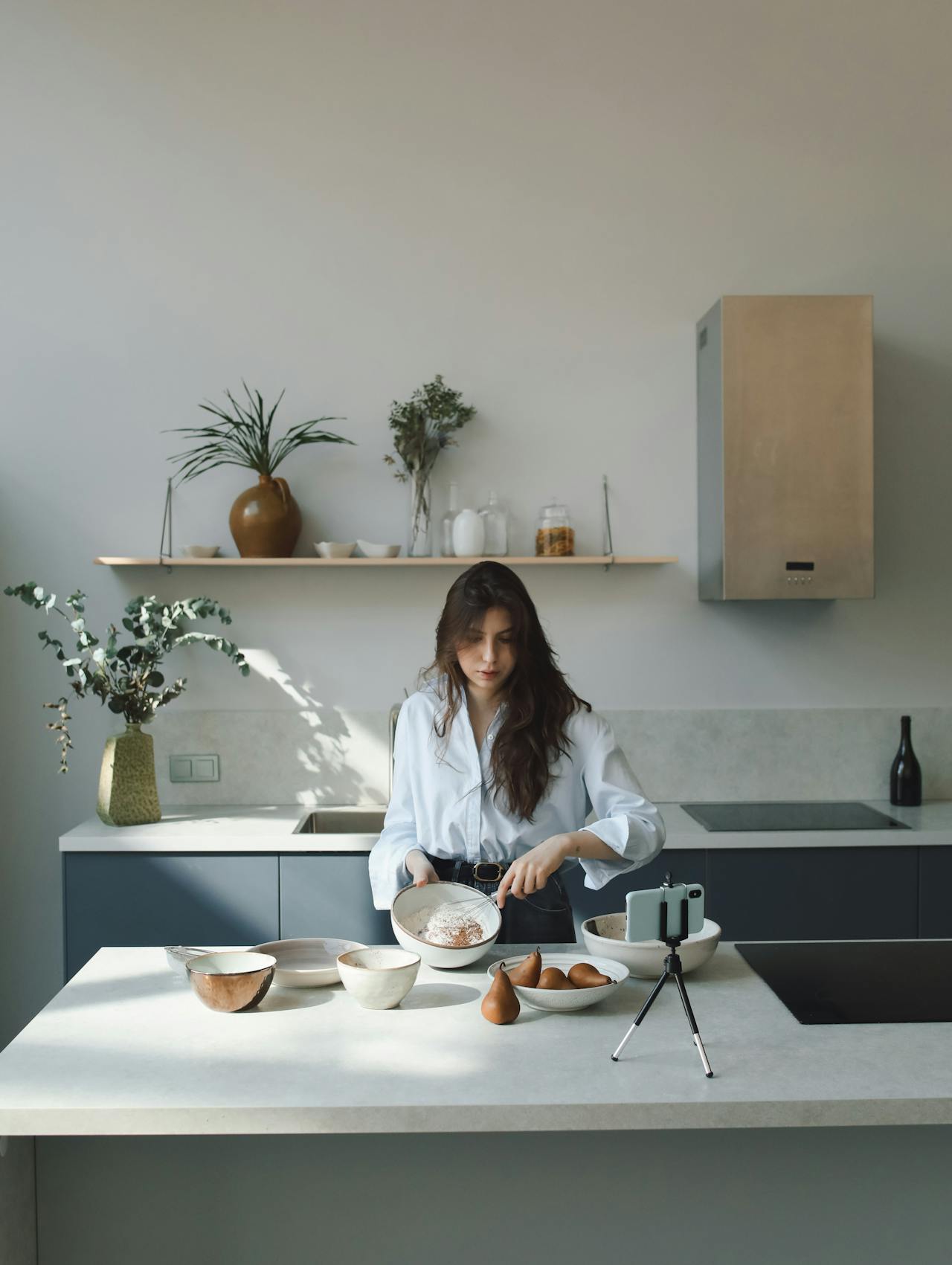 Image of a woman in a bright kitchen prepares food while filming herself with a smartphone on a tripod, representing self-care routine for moms.