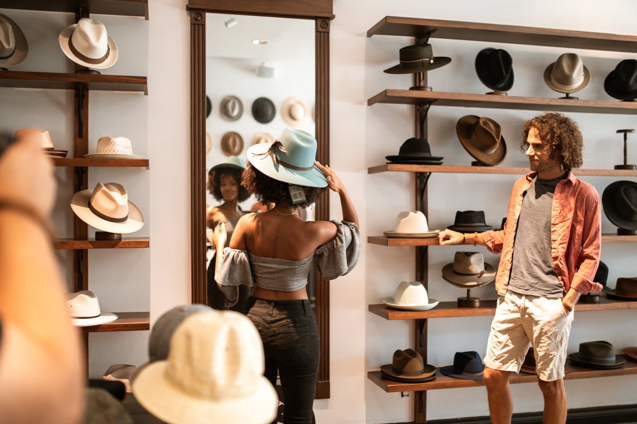 Image of a woman tries on a blue hat in a stylish hat store, looking at a mirror while a man observes nearby.