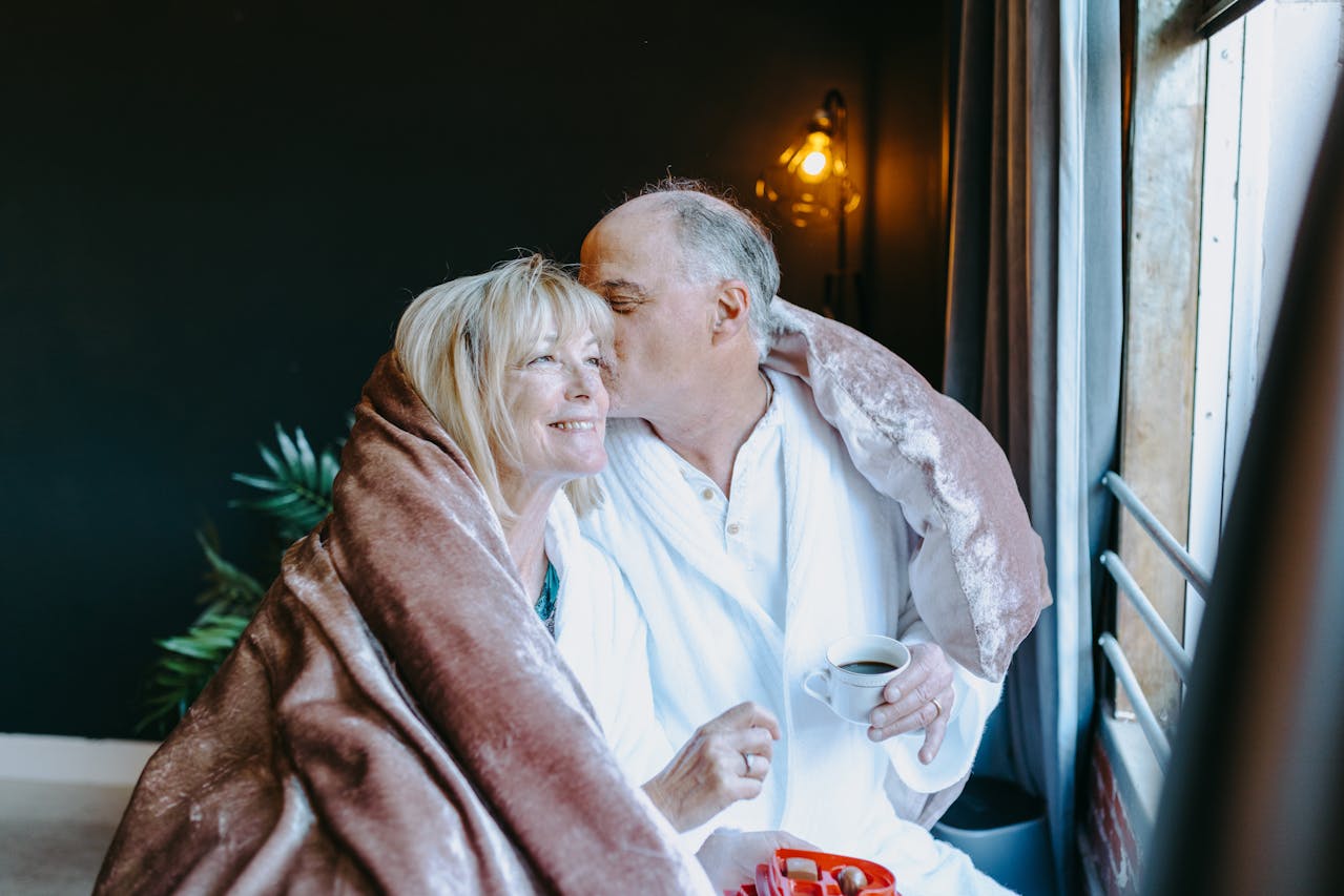 Image of a elderly couple shares a cozy moment by a window, wrapped in a soft blanket with coffee and snacks.