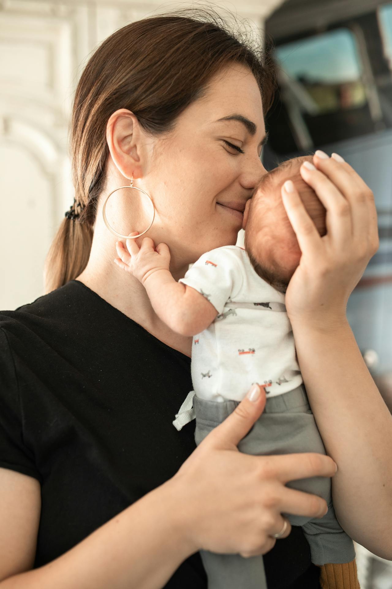 Image of a mother gently cradling and kissing her newborn baby, who is dressed in a white onesie with patterns, representing postpartum recovery before you start driving.