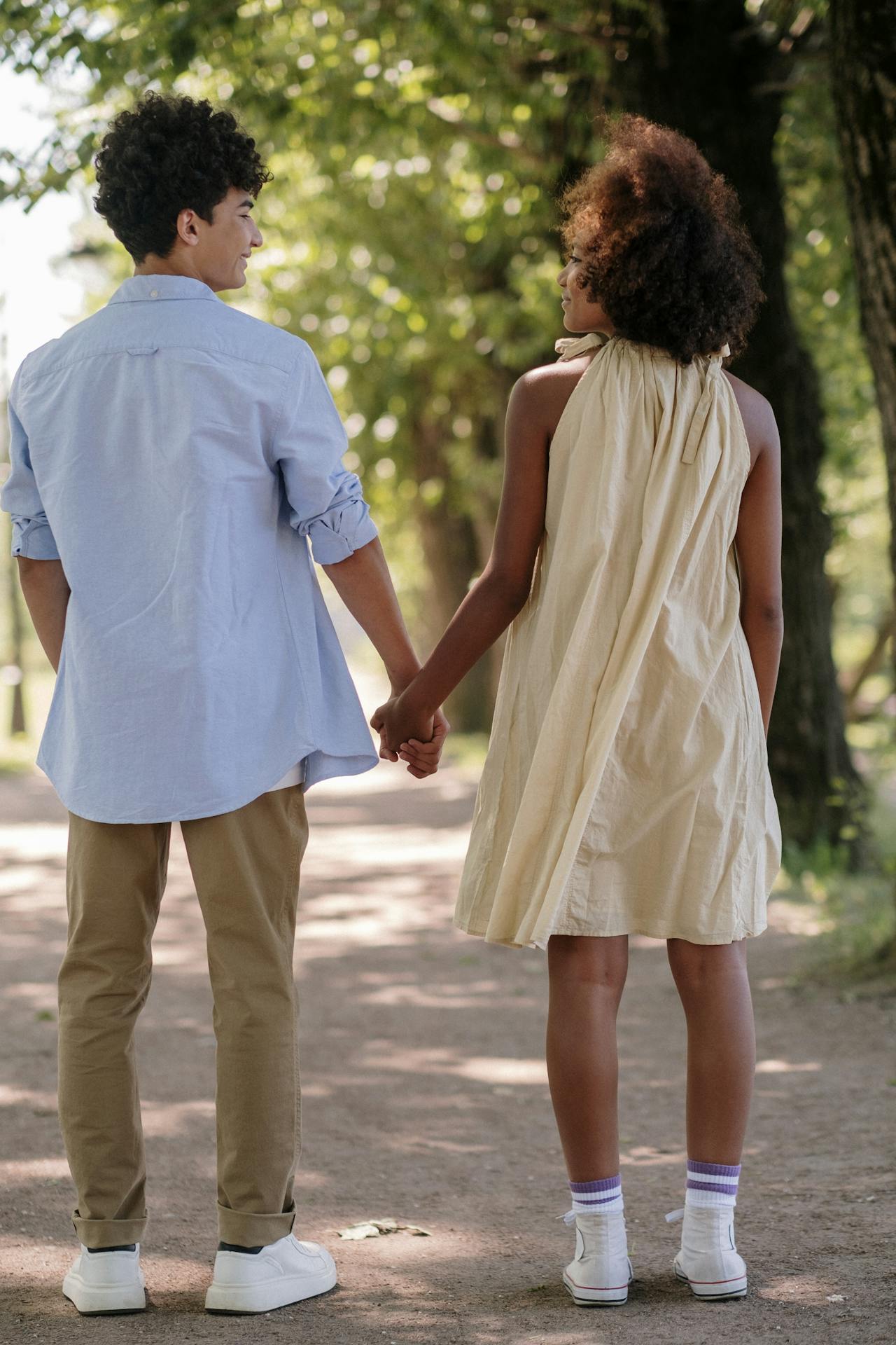 Image of a couple holding hands walks on a tree-lined path, the woman in a beige dress and the man in a blue shirt.