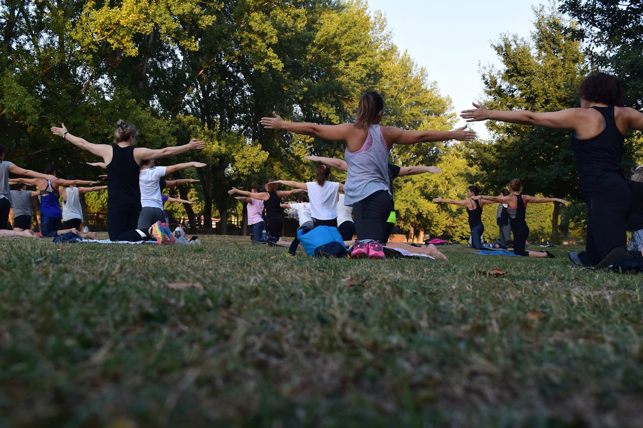 Image of a  group of people practicing outdoor yoga in a park, sitting on mats with arms outstretched in a pose.