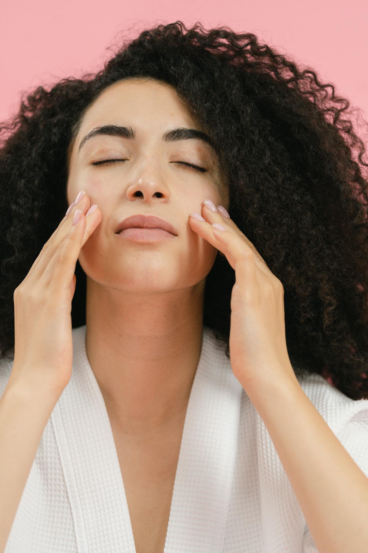 Image of a woman with curly hair massages her face with both hands while wearing a white robe against a pink background, showing self-care for a mom.