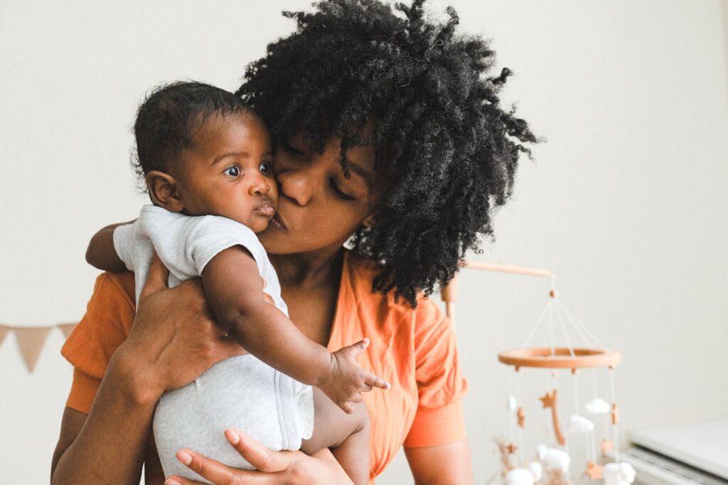 Close-up image of a mother in an orange top kisses her baby’s cheek in a softly lit nursery with a mobile in the background.