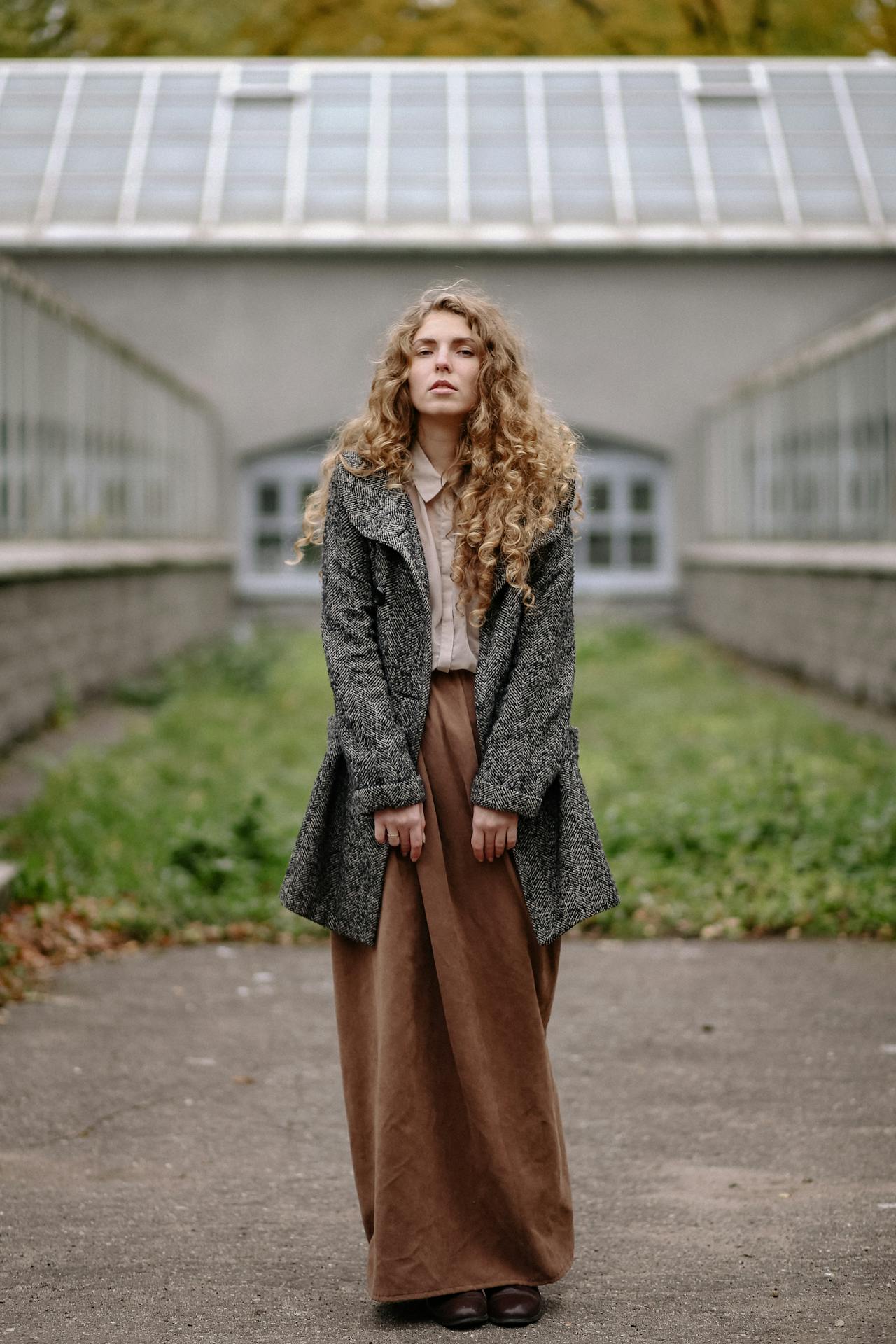 Image of a woman with curly hair standing outdoors, wearing a coat and long skirt in a neutral-colored setting, representing how can moms stay stylish and comfortable daily.