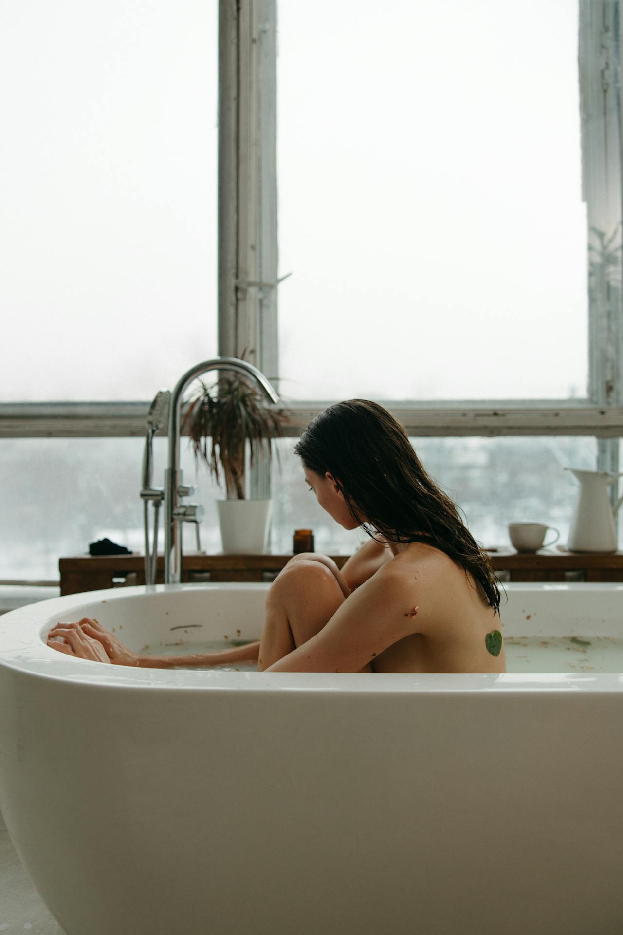 Image of a woman with wet hair sitting in a bathtub, looking out of a large window on a cloudy day, representing how to relieve postpartum itching.