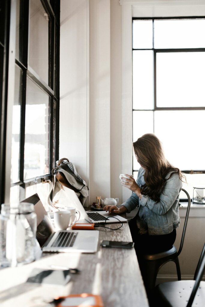 Image of a woman working at a window-side café desk using a laptop and holding a cup of coffee, showing how can a mother manage work-life balance.