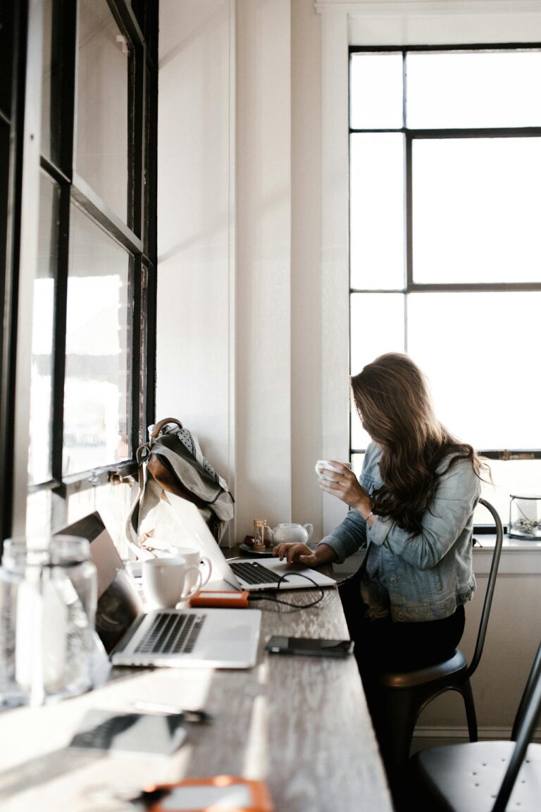 Image of a woman working at a window-side café desk using a laptop and holding a cup of coffee, showing how can a mother manage work-life balance.