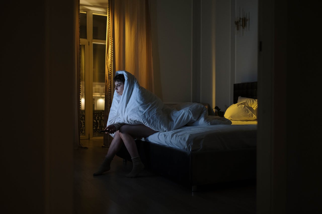 Why Mornings Are Challenging if You Have Depression? Image of a woman sitting on a bed at night wrapped in a blanket, watching TV in a dark room, showing why do I wake up feeling sad every day.