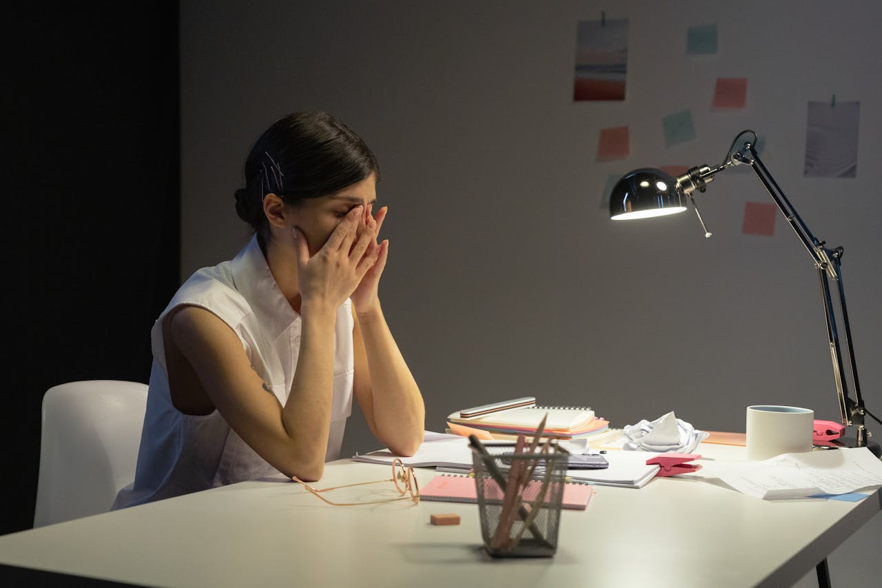 Why Mornings Are Challenging if You Have Depression? Image of a wired woman sitting at a cluttered desk, covering her face with her hands under a desk lamp.