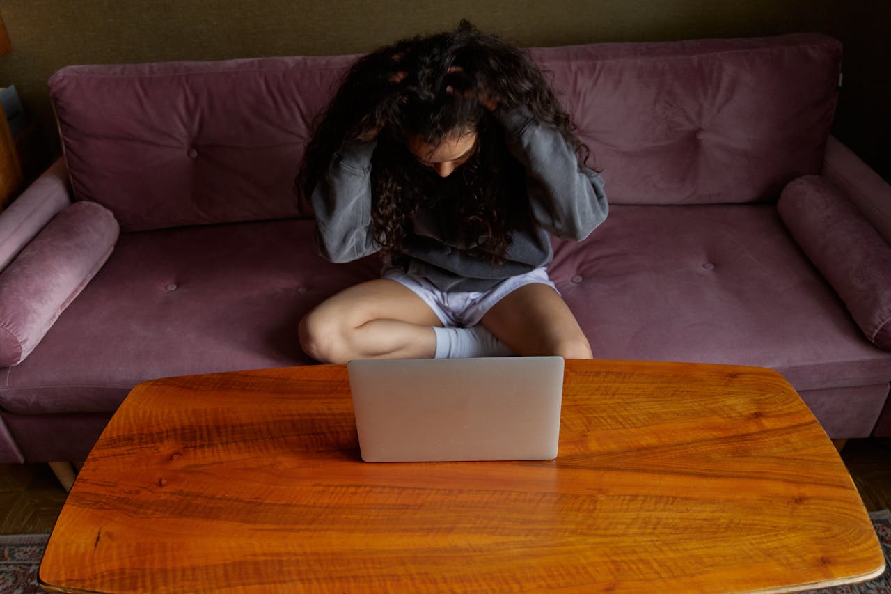 Why Mornings Are Challenging if You Have Depression? Image of a frustrated woman sitting cross-legged on a couch, grabbing her hair while looking at a laptop, representing why mornings are challenging if you have depression.
