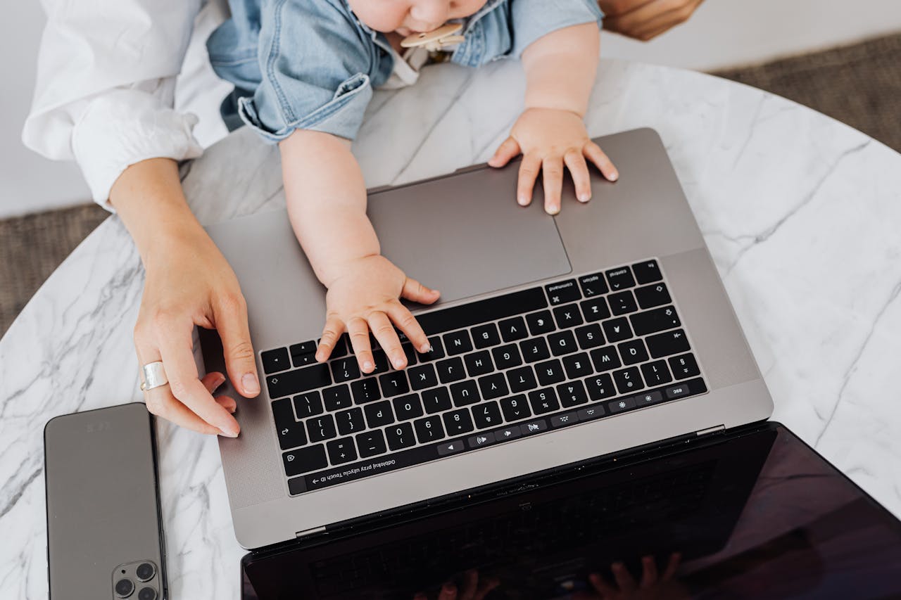How Can a Mother Manage Work-Life Balance? Image of a baby and adult hands typing on a laptop keyboard on a marble table with a smartphone nearby, representing how can a mother manage work-life balance.