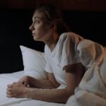 Image of a woman in a white t-shirt lying awake in bed at night, staring thoughtfully into the distance, showing why mornings are challenging if you have depression.